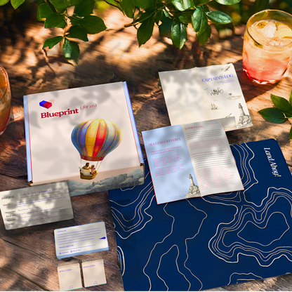 Collection of promotional materials including a book titled 'Blueprint' with a hot air balloon illustration, on a wooden surface with a natural background.