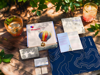 Outdoor setting with 'Blueprint' brochure, cards, and drinks on a wooden surface.
